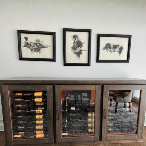 Wooden wine cabinet with glass doors displaying wine bottles, above which are three framed black and white artworks on a white wall.