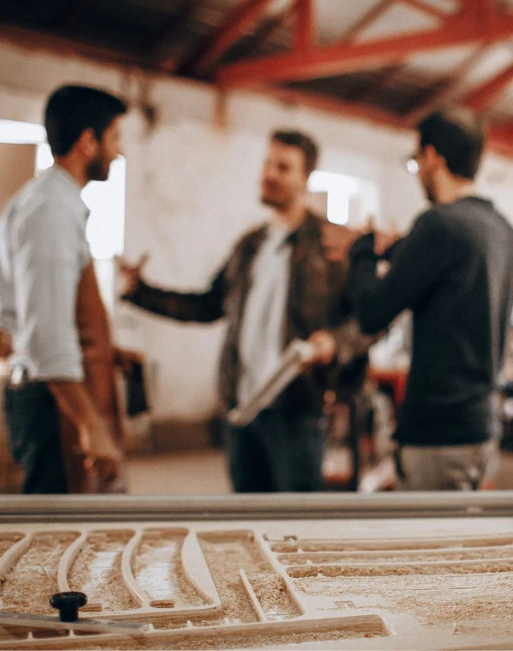 Three craftsmen collaborating in a woodworking shop with tools and wood shavings in the foreground.