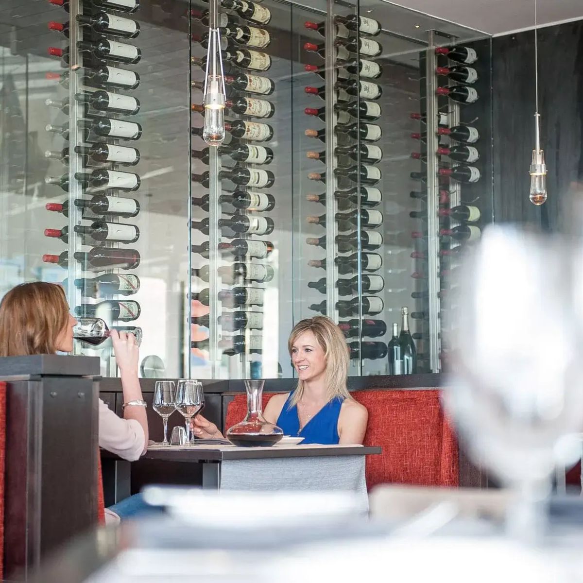 Two women sitting at a table in a wine cellar with wine bottles on the wall behind them.